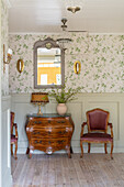 Antique chest of drawers and chair on landing with wood panelling and vintage-style wallpaper