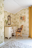 Dressing table and rattan chair in corner of room with floral wallpaper