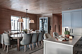 Oval dining table with grey upholstered chairs in elegant dining room with wood panelling and kitchen island in foreground