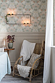 Rustic rattan armchair below modern candle sconce on wall with vintage-style wallpaper