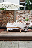 Gedeckter Tisch mit Sonnenschirm vor Backsteinmauer auf Holzterrasse