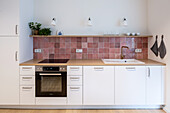 Kitchen counter with glazed tiled splashback in dusky pink below wall-mounted shelf
