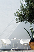 Weiße Schalenstühle und filigraner Tisch auf sonnigem Platz vor weißer Wand