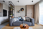 Grey upholstered sofa and coffee table in open-plan interior with shelves used as room divider and fitted kitchen
