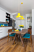 Weiße Einbauküche, Esstisch und Stühle mit blauen Sitzpolstern, darüber gelbe Pendelleuchten