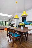 Dining table and chairs with blue seat cushions below yellow pendant lights in open-plan interior