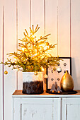 Christmas decoration with illuminated mini fir tree in a pot, golden vase and picture on chest of drawers