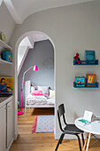 View through arched opening into a girl's bedroom with grey wall and pink accessories, in the foreground bookshelves and table