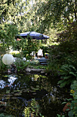 Lantern over a pond in a lush garden, in the background patio with parasol