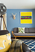 Wohnzimmer mit blauer Wand, gelbem Kunstwerk, dunkelgrauem Sofa und gemustertem Teppich