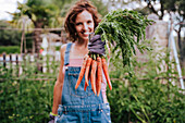 Lächelnde mittlere erwachsene Frau, die Karotten beim Stehen im Gemüsegarten hält