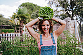 Lächelnde Frau, die Salat auf dem Kopf hält, während sie im Gemüsegarten steht