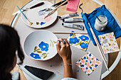 Crop talented female artist painting ceramic bowl while sitting at table with set of paintbrushes