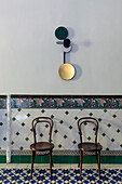 Zwei Thonet-Stühle vor Fliesenwand mit Bordüre im Sockelbereich, darüber Designer-Wandleuchte