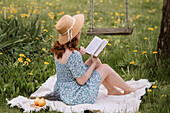 Rothaarige Frau im Kleid und Strohhut sitzt auf einer Picknickdecke und liest