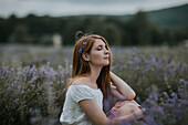Side view of gentle female with flowers in hair sitting in blooming lavender field and enjoying nature with closed eyes