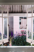Fenster mit Blick auf Blumengarten und rosa Holzhütte