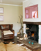 A traditional living room with a pink mirror above an open fireplace surrounded by logs and a leather arm chair next to a animal skin rug