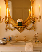 A detail of a modern bathroom a washbasin set in a cupboard unit an ornate gold mirror with candelabra
