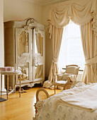 A traditional bedroom with a wooden floor an ornate antique wardrobe next to a window with swag curtains armchair
