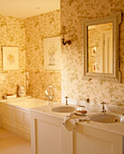 A traditional yellow bathroom with pattern wallpaper twin washbasins set in cupboard unit bathtub tap mirror