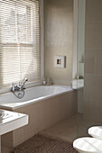 A modern bathroom with a window with Venetian blinds overlooking the bathtub surrounded by beige tiles