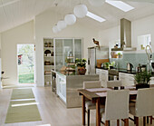 Eine moderne offene Wohnküche im Landhausstil mit Esstisch und Stühlen aus Holz, weißen Küchenschränken und einer offenen Tür nach draußen