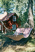 An outside detail of a striped hammock tied between trees in a garden wooden country house in the background