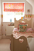 A detail of a children's room decorated in pink colours single bed with decorative wooden headboard roman blind at window floral pattern bed cover and wallpaper