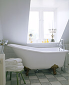 A traditional bathroom freestanding bathtub below a sloping ceiling with a large window and a tiled floor