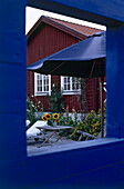 Blick durch ein quadratisches blaues Fenster auf ein Holzhaus