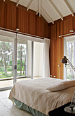Modernes, mit Zedernholz verkleidetes Schlafzimmer mit Betonboden und Glasschiebetüren zum Balkon mit Blick auf den Waldgarten