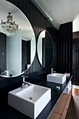 Double basins below circular mirrors in black panelled bathroom with chandelier light fitting