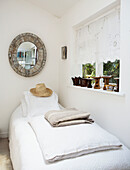 Sunhat on single bed below window in white bedroom with circular mirror