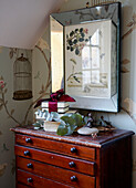 Wooden chest of drawers under artwork with floral patterned wallpaper