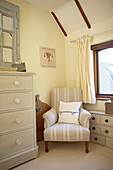 Gingham checked curtains at window of cottage bedroom with beige armchair and painted unit