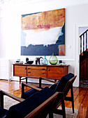 Wooden sideboard below artwork in living room with matching blue upholstered chairs