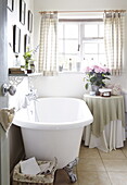 Freestanding bath with shelf and artwork at window in farmhouse cottage, Oxfordshire, England, UK
