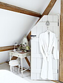 White bathrobe hanging on back of attic bedroom door next to dressing table and stool, Oxfordshire, England, UK