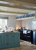 Blick auf den schwarzen Aga-Elektroherd in der Landhausküche mit hellblau gestrichener Kücheninsel mit Holzarbeitsplatte und einem Regal mit blauen und weißen Dekotellern