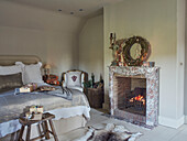 Wreath above lit fire with gifts wrapped in brown paper on silver bed cover in Oxfordshire bedroom England UK