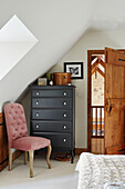 Pink buttoned chair with black tallboy and wooden bedroom door in Kent home, North East England, UK