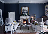 Pair of sofas with grey ottoman and lit fire in blue East Grinstead living room, West Sussex, UK