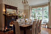 Slip-covered dining chairs with lit candles and in dining room with antique fireplace and bay window, UK home