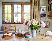 Cut peonies and sponge cake on table with leaded glass window in Sandford St Martin cottage, Oxfordshire, UK
