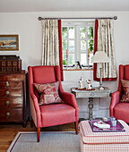 Pair of red armchairs and wooden chest of drawers at window in Cotswolds cottage, UK