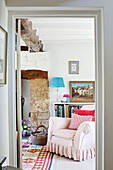 Pastel pink armchair at exposed stone fireside with bookcase in, UK cottage