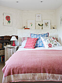 Patterned cushions and framed botanic prints with pink blanket in whitewashed cottage bedroom, UK