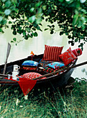 Ruderboot mit bunten Kissen, Decken und Flachmann an einem Teich in einem Garten