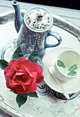 Detail einer Teekanne und einer dekorativen roten Rose auf einem Tablett auf einer Tischplatte